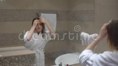 穿着白色长袍的有趣迷人的女孩洗了澡，用吹风机在镜子前唱歌。